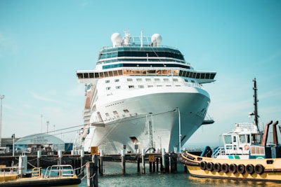 A large cruise ship is docked at a port, with a smaller tugboat nearby. The ship is prominent in the center, displaying a sleek white design and multiple decks. The sky above is clear and blue, creating a vibrant backdrop.