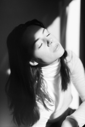 A person with long hair is wearing a light-colored turtleneck and appears to be in a state of tranquility, with eyes closed and face tilted toward the light. Shadows create a dramatic contrast across the face.