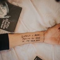 A forearm with the written text 'LATELY I BEEN FEELING SO DEAD IN MY OWN SKIN' and a partial view of the book 'Fifty Shades Darker' against a light fabric background.