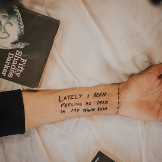 A forearm with the written text 'LATELY I BEEN FEELING SO DEAD IN MY OWN SKIN' and a partial view of the book 'Fifty Shades Darker' against a light fabric background.