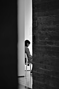 A black and white image of a person sitting on a chair, viewed through a slightly open door. The scene is mostly in shadow, with the person partially obscured by a light wall and the door. The setting appears quiet and contemplative.