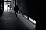 A person is seen walking in a dimly lit hallway with a broom in hand, near a large glass exit door. Light enters from the door and window, casting shadows on the tiled floor.