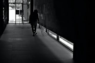 A person is seen walking in a dimly lit hallway with a broom in hand, near a large glass exit door. Light enters from the door and window, casting shadows on the tiled floor.