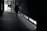 A person is seen walking in a dimly lit hallway with a broom in hand, near a large glass exit door. Light enters from the door and window, casting shadows on the tiled floor.