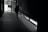 A person is seen walking in a dimly lit hallway with a broom in hand, near a large glass exit door. Light enters from the door and window, casting shadows on the tiled floor.