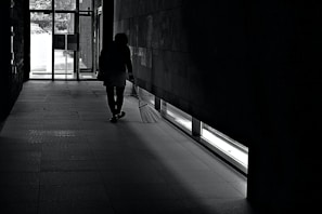 A person is seen walking in a dimly lit hallway with a broom in hand, near a large glass exit door. Light enters from the door and window, casting shadows on the tiled floor.