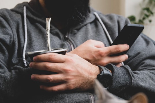 A person with a beard is holding a mate cup in one hand and a smartphone in the other. The individual is wearing a dark gray hoodie and a black wristwatch. The scene suggests a relaxed moment, possibly at home.