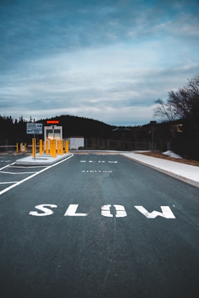 A road with the word 'SLOW' painted in large white letters. The sky is overcast with a hint of blue. On the left side, there are parking spaces marked, and yellow bollards line the entrance to a small booth in the background, labeled 'ENTRANCE'. Trees and a fence line the horizon.