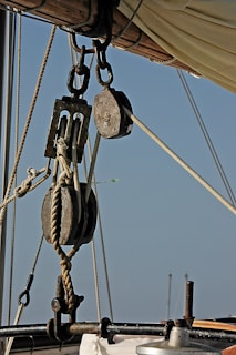 A close-up view of wooden pulleys and rigging on a sailboat, with ropes intricately looped through them. The background shows a clear blue sky, highlighting the nautical hardware and sails visible above.