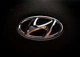 A chrome Hyundai logo with a sleek, rounded design is set against a textured black background, typically found on a steering wheel or vehicle interior.