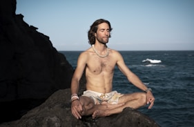 topless man in brown shorts sitting on rock near sea during daytime