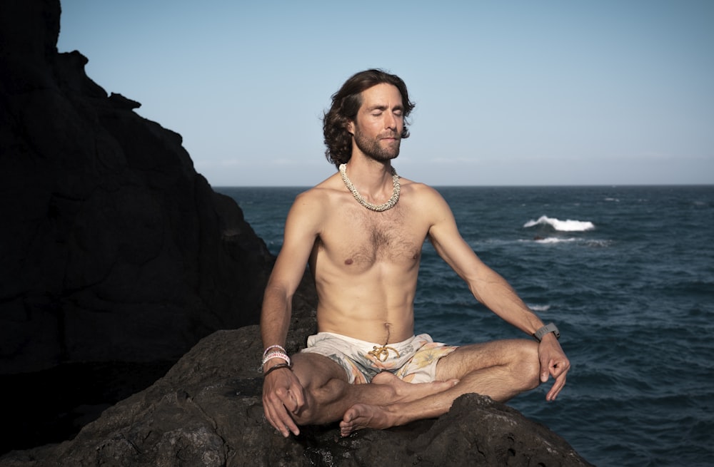 uomo in topless in pantaloncini marroni seduto sulla roccia vicino al mare durante il giorno