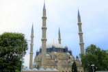 A grand mosque with multiple tall, slender minarets and a large central dome is surrounded by lush green trees. The architectural style is classic Islamic, featuring intricate stonework and arches.
