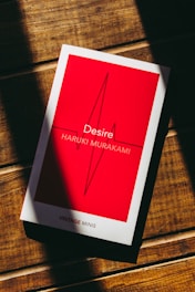 A book with a red cover and white border lies on a wooden surface. The title 'Desire' and author 'Haruki Murakami' are printed on the cover, along with an abstract line design. Light and shadow create stripes across the surface.