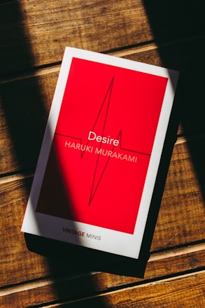 A book with a red cover and white border lies on a wooden surface. The title 'Desire' and author 'Haruki Murakami' are printed on the cover, along with an abstract line design. Light and shadow create stripes across the surface.
