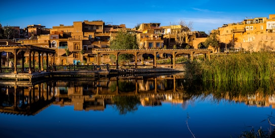 A landscape featuring traditional buildings in warm earth tones reflected in a calm body of water. The scene includes a series of interconnected wooden structures resembling gazebos, with lush greenery in the foreground. Clear blue sky and calm waters enhance the overall tranquility.
