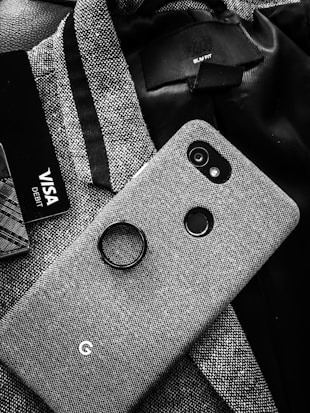 A textured fabric phone case is positioned on top of a matching fabric pattern. Beside it, there is a partial view of a Visa debit card, and the label of a slim-fit jacket is visible in the top-right corner. The monochrome composition enhances the detailed patterns of the fabric.