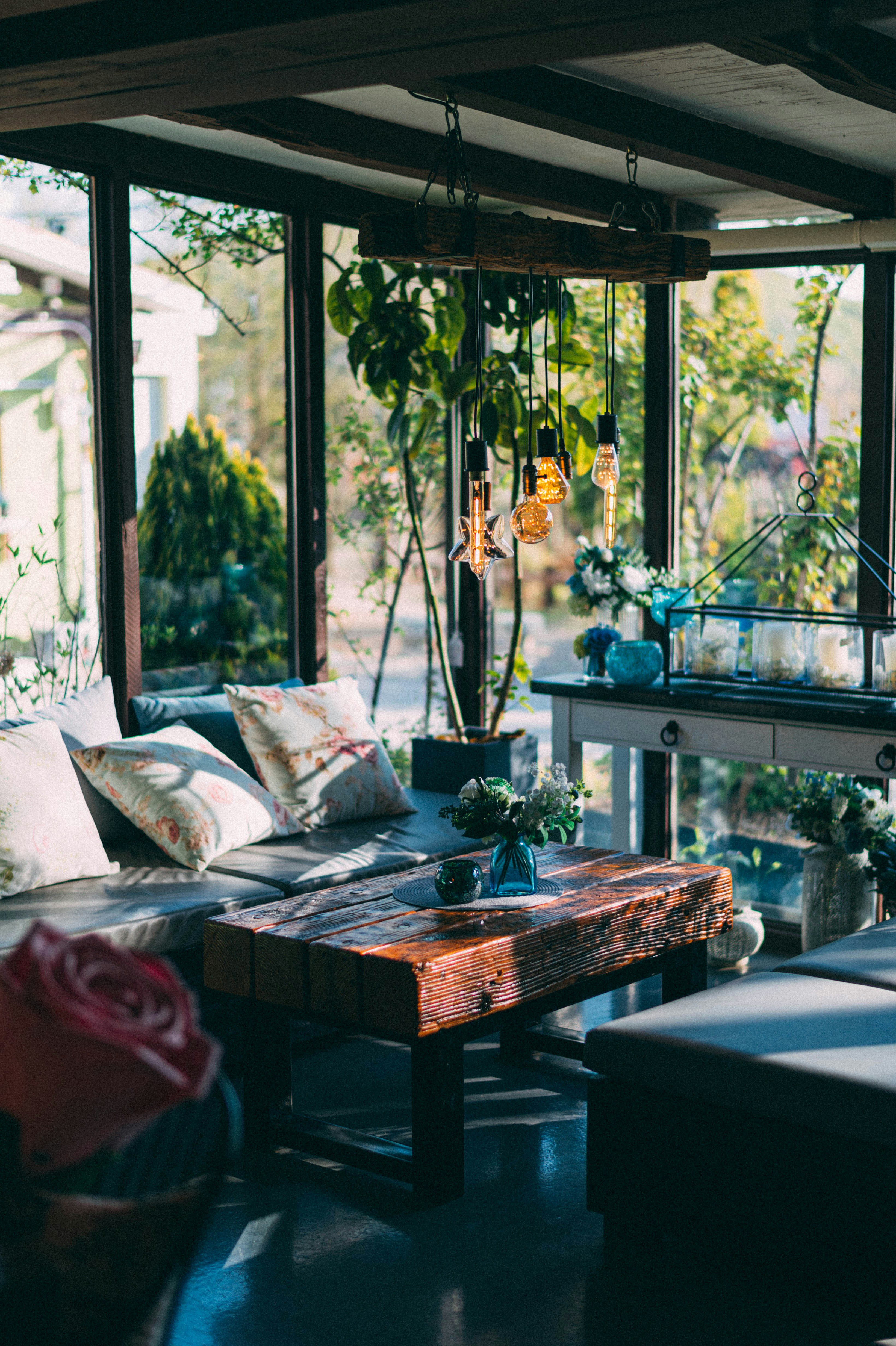 Bright conservatory with large windows, cushioned seating, rustic wooden table, hanging lights, and potted plants.