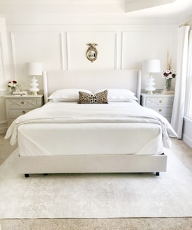 A neatly arranged bedroom with a large bed centered against a white panelled wall featuring a decorative gold-framed mirror. Beside the bed are two matching nightstands with geometric designs, topped with white lamps and vases of flowers. A leopard print pillow is placed in the middle of the bed.