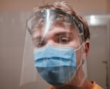 A young person is wearing a blue surgical mask and a clear face shield, looking directly at the camera. The background is out of focus, suggesting an indoor setting.