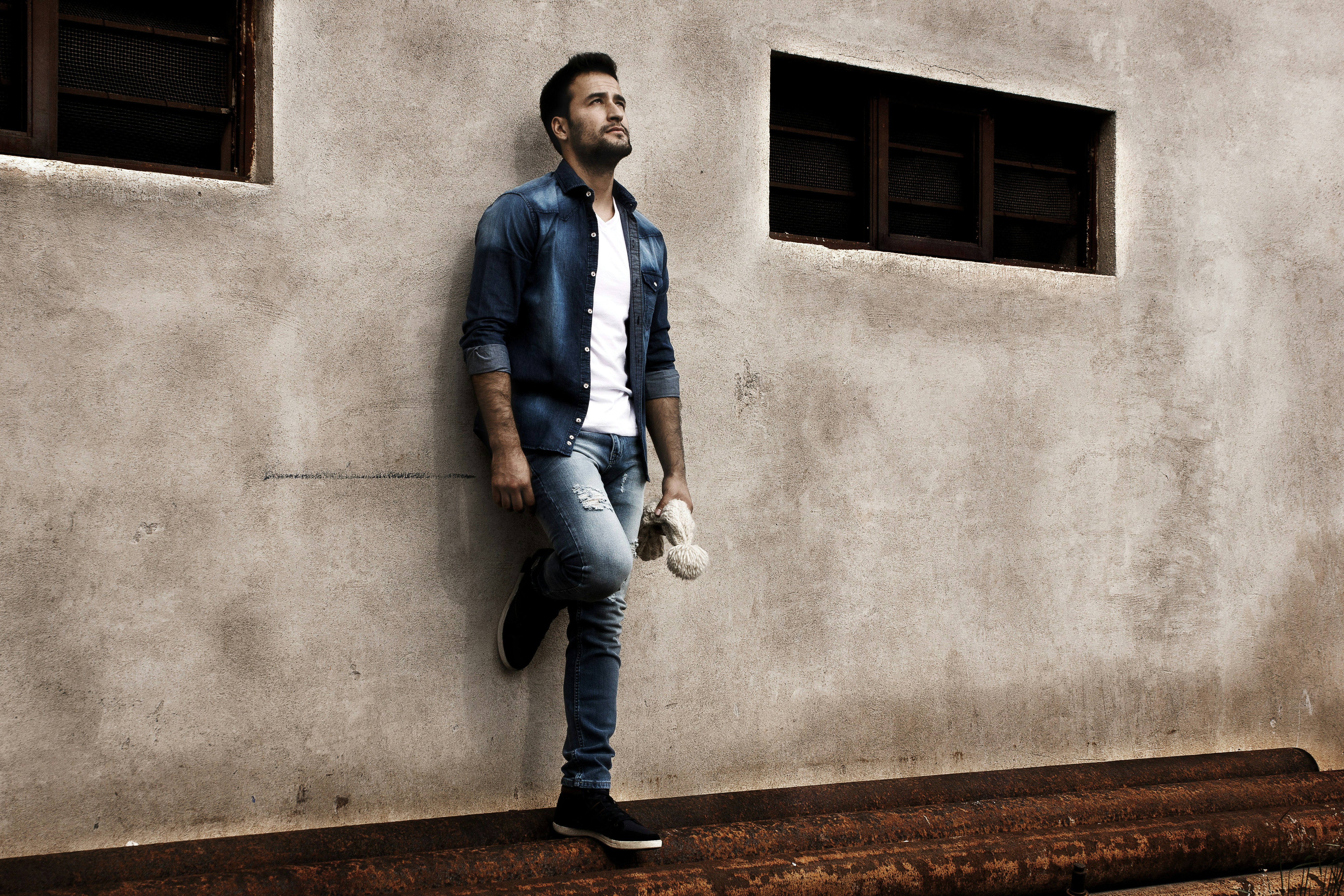 man in blue dress shirt and blue denim jeans standing beside gray concrete wall during daytime