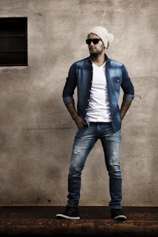 man in blue blazer and blue denim jeans standing beside gray wall