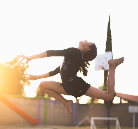 A young gymnast is captured in mid-air during a graceful leap, with her arms and legs extended. The golden sunlight creates a warm glow around her, highlighting her movements. In the background, a basketball hoop and playground equipment are visible, suggesting an outdoor setting.