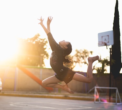 A person gracefully leaps into the air against a sunlit background, showcasing a gymnastics or dance move. The surrounding environment includes a basketball hoop and outdoor sports court.