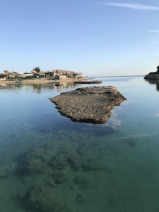 A peaceful coastal landscape with calm, clear water reflecting the blue sky. Large rock formations are visible, surrounded by gentle ripples. In the background, there are buildings with red-tiled roofs and palm trees. The scene conveys a sense of tranquility and natural beauty.