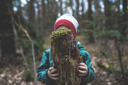 child in blue jacket and white knit cap holding brown tree trunk