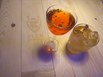 Two elegantly presented cocktails sit on a wooden table. The cocktail on the left is in a coupe glass filled with a vibrant orange liquid adorned with small green garnish elements. The cocktail on the right, placed in an old-fashioned glass, is a pale yellow drink topped with slices of a dried fruit and ice cubes. The table features a faint engraving of a deer emblem with the text 'VI.ER DANMARK' beside the drinks.