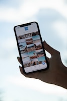 A hand holds a smartphone displaying a gallery view of images. The phone screen shows various photos in a grid layout, featuring landscapes, architecture, and possibly travel destinations. The background is a blurred blue sky, suggesting a serene or outdoor setting.