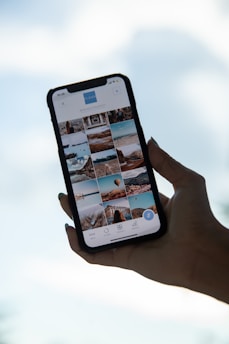 A hand holds a smartphone displaying a gallery view of images. The phone screen shows various photos in a grid layout, featuring landscapes, architecture, and possibly travel destinations. The background is a blurred blue sky, suggesting a serene or outdoor setting.