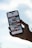 A hand holds a smartphone displaying a gallery view of images. The phone screen shows various photos in a grid layout, featuring landscapes, architecture, and possibly travel destinations. The background is a blurred blue sky, suggesting a serene or outdoor setting.