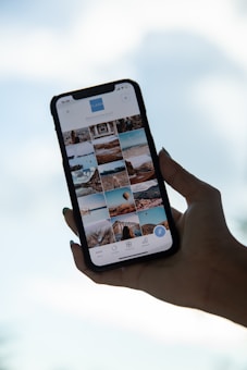 A hand holds a smartphone displaying a gallery view of images. The phone screen shows various photos in a grid layout, featuring landscapes, architecture, and possibly travel destinations. The background is a blurred blue sky, suggesting a serene or outdoor setting.
