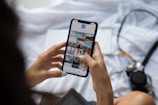Someone is holding a smartphone and browsing a photo gallery app. The device displays several thumbnail images featuring various landscapes and architectural scenes. The person has manicured nails painted in a light blue color. A blurred camera and a keyboard can be seen in the background on a light-colored fabric surface.