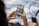 Someone is holding a smartphone and browsing a photo gallery app. The device displays several thumbnail images featuring various landscapes and architectural scenes. The person has manicured nails painted in a light blue color. A blurred camera and a keyboard can be seen in the background on a light-colored fabric surface.