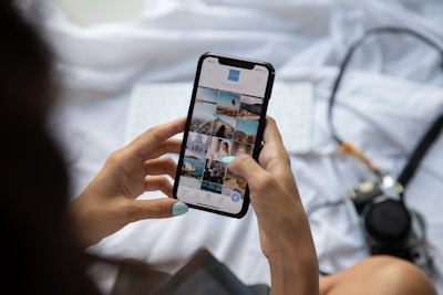 Someone is holding a smartphone and browsing a photo gallery app. The device displays several thumbnail images featuring various landscapes and architectural scenes. The person has manicured nails painted in a light blue color. A blurred camera and a keyboard can be seen in the background on a light-colored fabric surface.