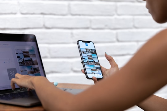 A person is using a smartphone and a laptop simultaneously. The smartphone screen shows a grid of photos, possibly a photo gallery or social media app. The laptop screen displays a similar photo interface. Both devices are being used casually, possibly to edit or upload pictures. The background is a white, textured brick wall.