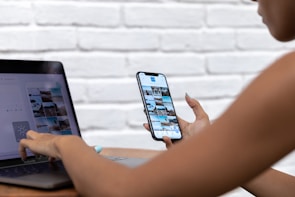 A person is using a smartphone and a laptop simultaneously. The smartphone screen shows a grid of photos, possibly a photo gallery or social media app. The laptop screen displays a similar photo interface. Both devices are being used casually, possibly to edit or upload pictures. The background is a white, textured brick wall.