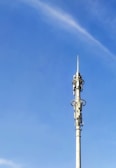 A tall communication tower with various antennas and dishes, set against a clear blue sky. The tower has multiple platforms and technical equipment attached at intervals along its height.
