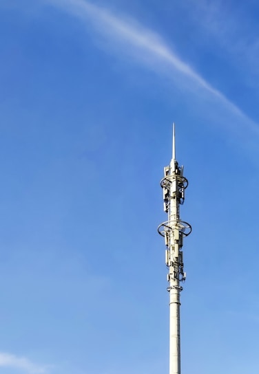 A tall communication tower with various antennas and dishes, set against a clear blue sky. The tower has multiple platforms and technical equipment attached at intervals along its height.