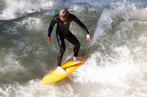 A person wearing a black wetsuit is surfing on a vibrant yellow surfboard amid white, foamy waves. The action captures a dynamic display of balance and control on the water.