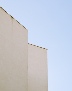 Minimalist architectural composition featuring a beige building contrasted against a clear blue sky. The structure has clean lines and flat surfaces, creating a sense of simplicity and calm.