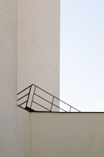 A minimalistic architectural image featuring a beige concrete structure with clean lines and a simple design. An angled black metal railing creates a geometric pattern against the wall, contrasting with the stark, pale background.