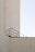 A minimalistic architectural image featuring a beige concrete structure with clean lines and a simple design. An angled black metal railing creates a geometric pattern against the wall, contrasting with the stark, pale background.