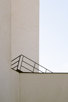 A minimalistic architectural image featuring a beige concrete structure with clean lines and a simple design. An angled black metal railing creates a geometric pattern against the wall, contrasting with the stark, pale background.