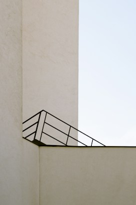 A minimalistic architectural image featuring a beige concrete structure with clean lines and a simple design. An angled black metal railing creates a geometric pattern against the wall, contrasting with the stark, pale background.