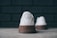 A white sneaker is positioned on a concrete surface with the focus on the heel. The sneaker has a brown textured sole and a stitched logo on the back. The background features a subtle, out-of-focus dark brick wall.