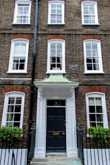 A traditional brick townhouse with three floors and symmetrical architecture. The facade features multiple white-framed windows and a prominent black front door with a white portico. Iron railings and window boxes with greenery and red flowers are present.